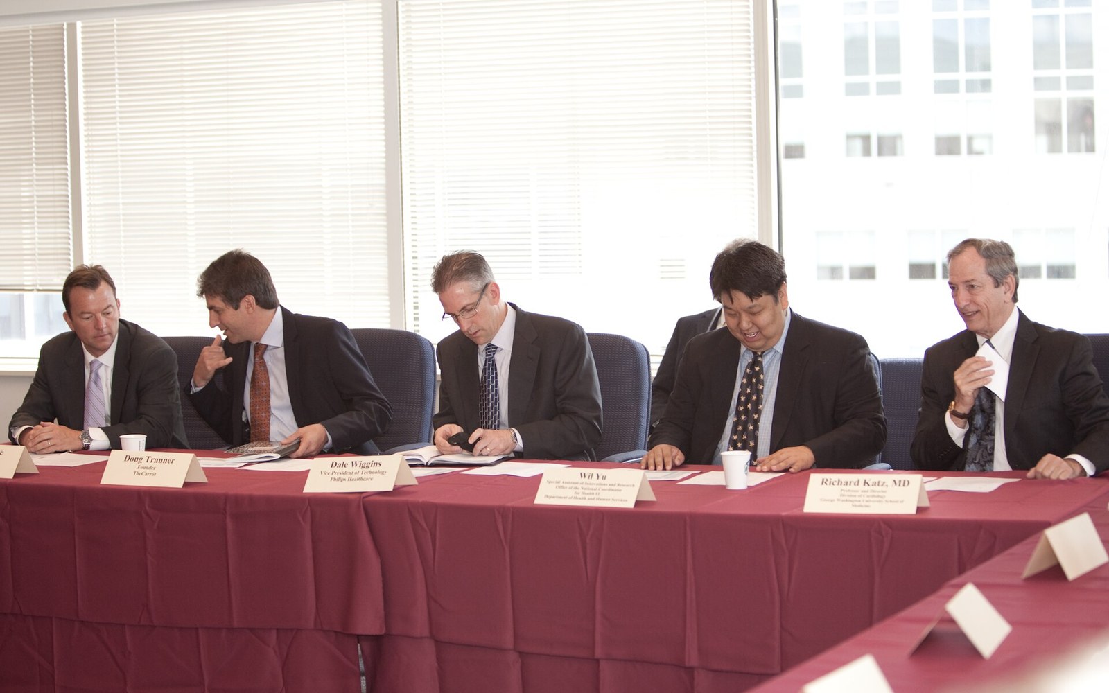 Five panelists in suits sit behind name placards at a maroon-clothed conference table