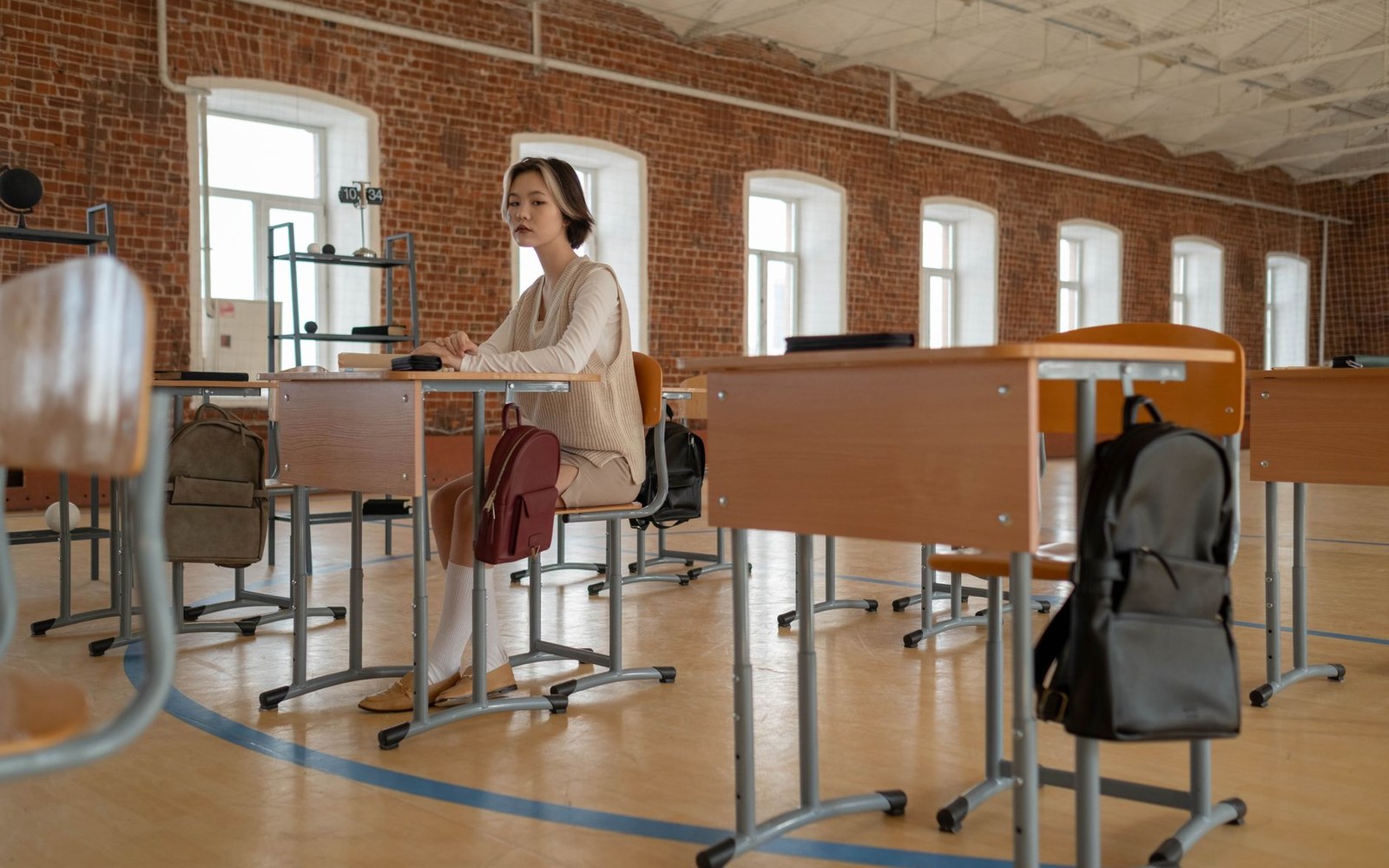 A woman sits alone at a desk among rows of empty desks with backpacks hanging on chairs