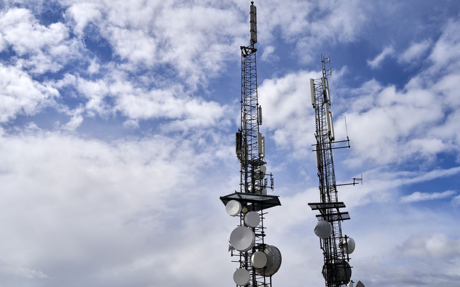 Two telecommunications towers with satellite dishes and antennas against a partly cloudy sky
