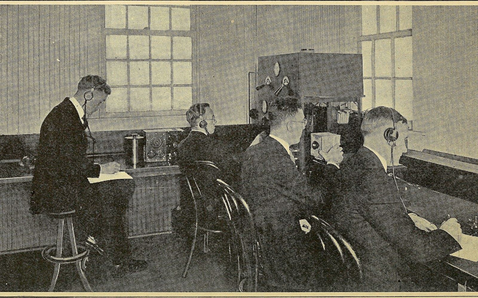 Men wearing headphones operate early radio receiving equipment at a row of desks