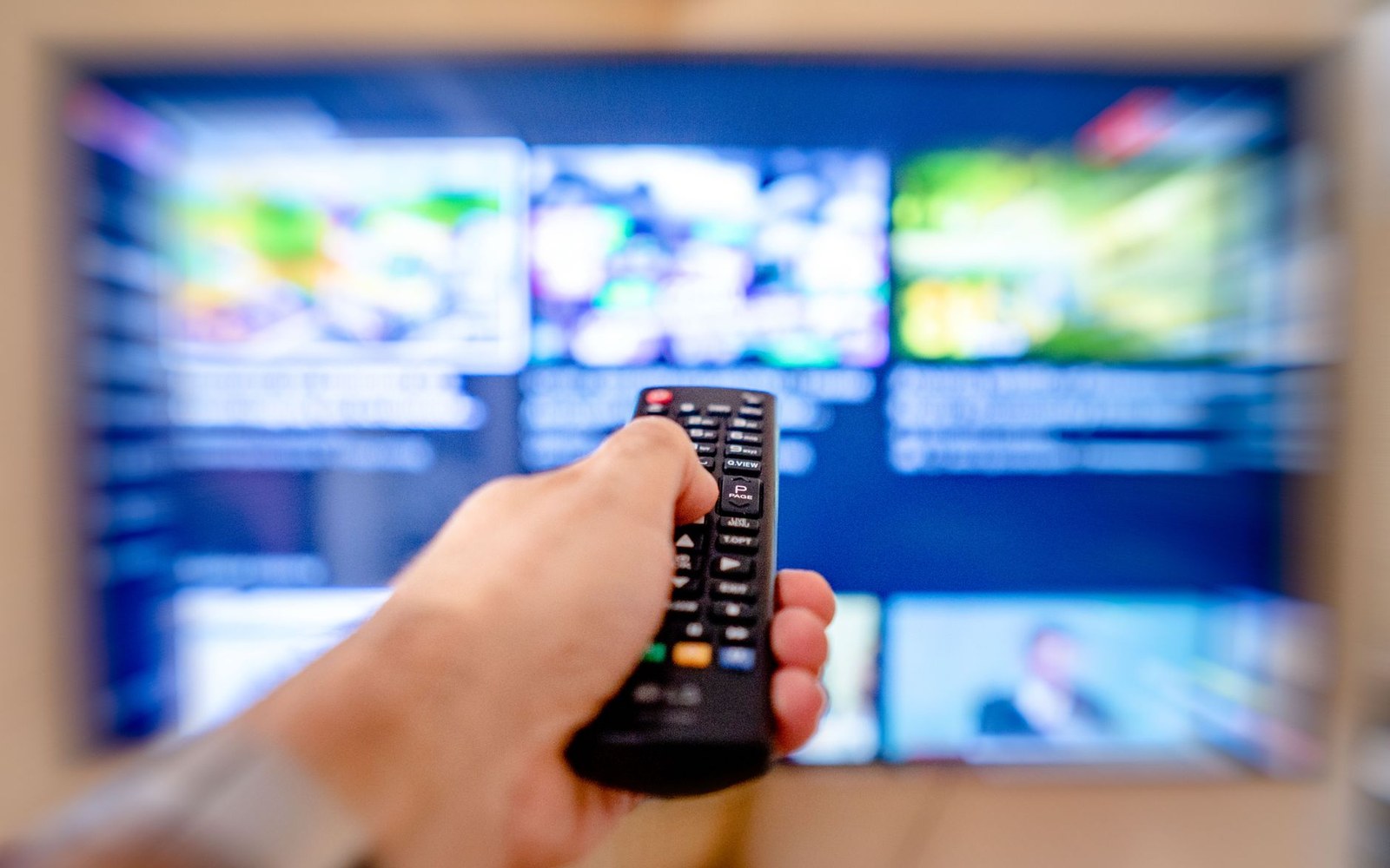 A hand points a remote control toward a blurred grid of television programming thumbnails