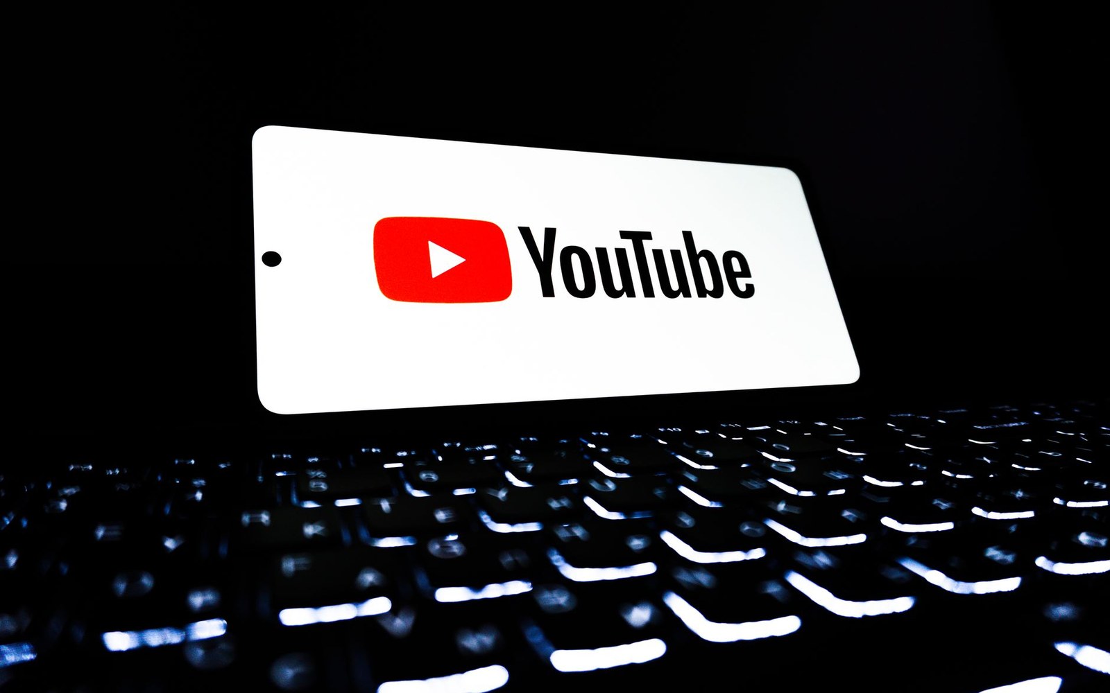 Smartphone displaying the YouTube logo propped above an illuminated keyboard