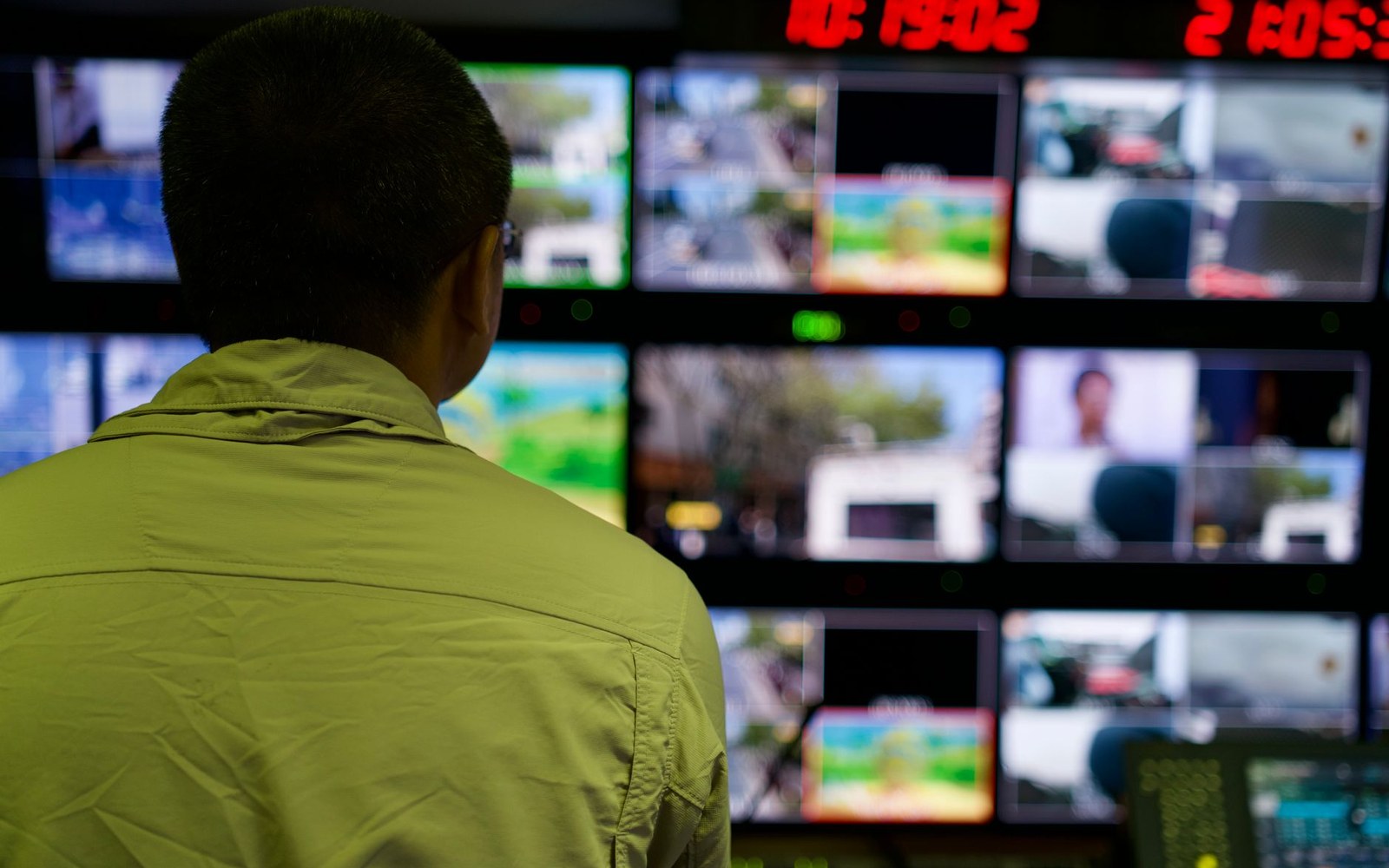 A television gallery control room mid-broadcast, an adult director at the desk facing a wall of preview monitors showing a live news programme